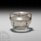 Roman Iridescent Handled Glass Vessel. 3rd-4th century A.D. A clear glass vessel with carinated body, everted neck, three applied trails around the sh...