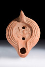 Ca. 100-300 AD.
A terracotta oil lamp with a flattened base, smooth side walls, grooved perimeter, and a recessed tondo decorated with a hippocampus -...