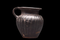 Ca. 400-300 BC.
A black glazed mug featuring an elegant inverted pear-shaped body with incised vertical grooves around the body. The mug has a short c...