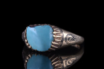 Persia, Ca 12th century AD.
A nice silver ring with a round hoop, and expanding shoulders decorated with niello motifs. A large bezel set with turquoi...
