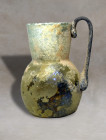 Iridescent light blue blown glass bottle with single handle. The handle is a deep dark colour. It has iridescence and parts with earthy traces. Invent...