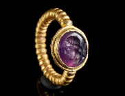 A LATE ROMAN GOLD RING WITH AN AMETHYST INTAGLIO Circa 4th century AD. Composed of a solid hoop formed from beaded gold wire and an offset oval bezel....