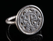 A LATE MEDIEVAL SILVER RING WITH A FLEUR-DE-LIS Circa 15th century AD. Large circular bezel mounted on a twisted hoop. The bezel engraved with a Fleur...