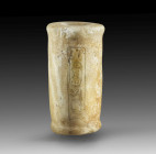 Cylindrical alabaster mock vessel with a stepped bulge on the lip and base. The interior is only partially drilled. The exterior is incised with a car...