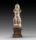 Light red terracotta figurine of Isis-Aphrodite depicted as a nude woman balancing a basket on her head with both raised hands. A remnant of a garment...