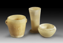 Three alabaster vessels. New Kingdom - Late Period, 1550 - 332 BC. a) Mug with two lugged handles. H 5.8cm. Rim slightly chipped, otherwise intact. b)...