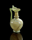Juglet of clear green glass with thumb-rest. H 19cm. Globular body with a ring foot, long neck and trefoil mouth with folded rim; the handle folded on...