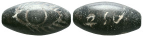 This is an elongated, oval-shaped ancient seal or amulet made from dark stone, possibly serpentine or basalt. The surface of the object shows engraved...